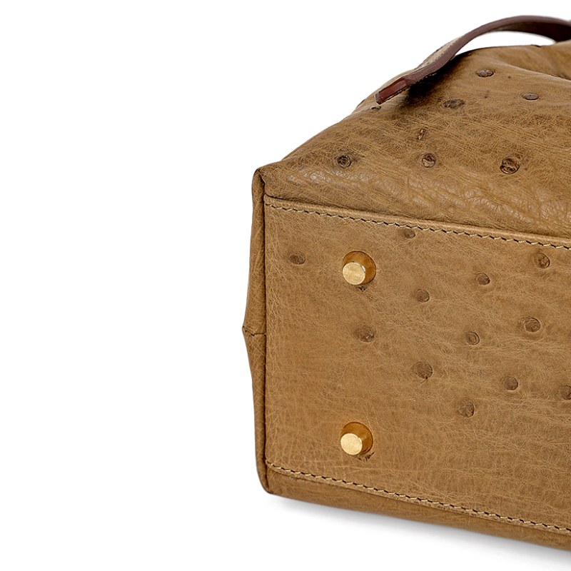 Close-up of a tan leather bag corner with visible stitching and gold studs on an ostrich-textured surface