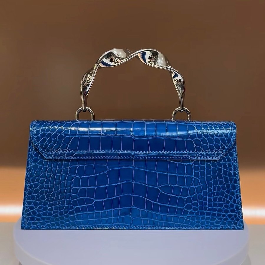 Blue crocodile-textured handbag with a twisted silver metal handle on a display pedestal.